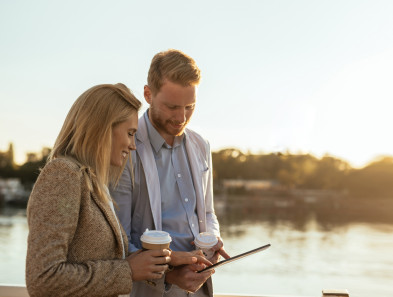 Two people walking along water while looking at a device and holding hot drinks.