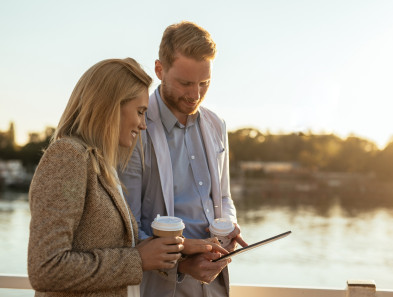 Two people walking along water while looking at a device and holding hot drinks.