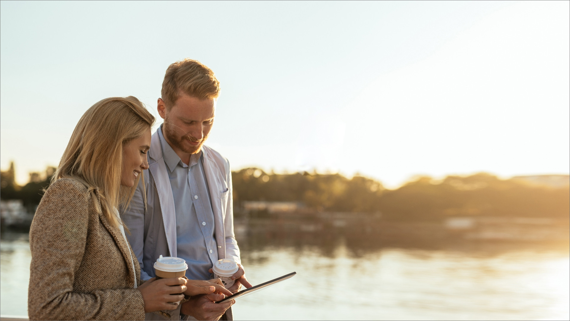 Two people walking along water while looking at a device and holding hot drinks.