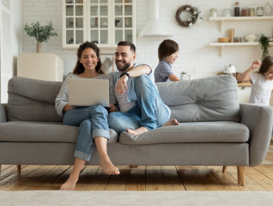 A couple looking at a laptop together while sitting on a couch, with kids playing around them